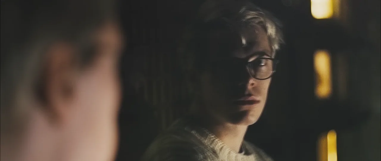 Still from Insanity: a young man in glasses stares past an out-of-focus figure in a dim, lamplit library.