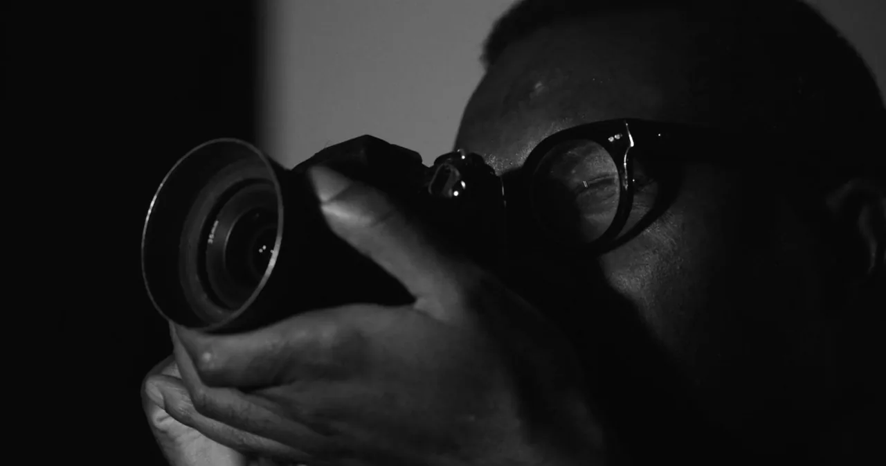Black and white close-up from The Trace: a street photographer in glasses raises his camera, lens towards us.