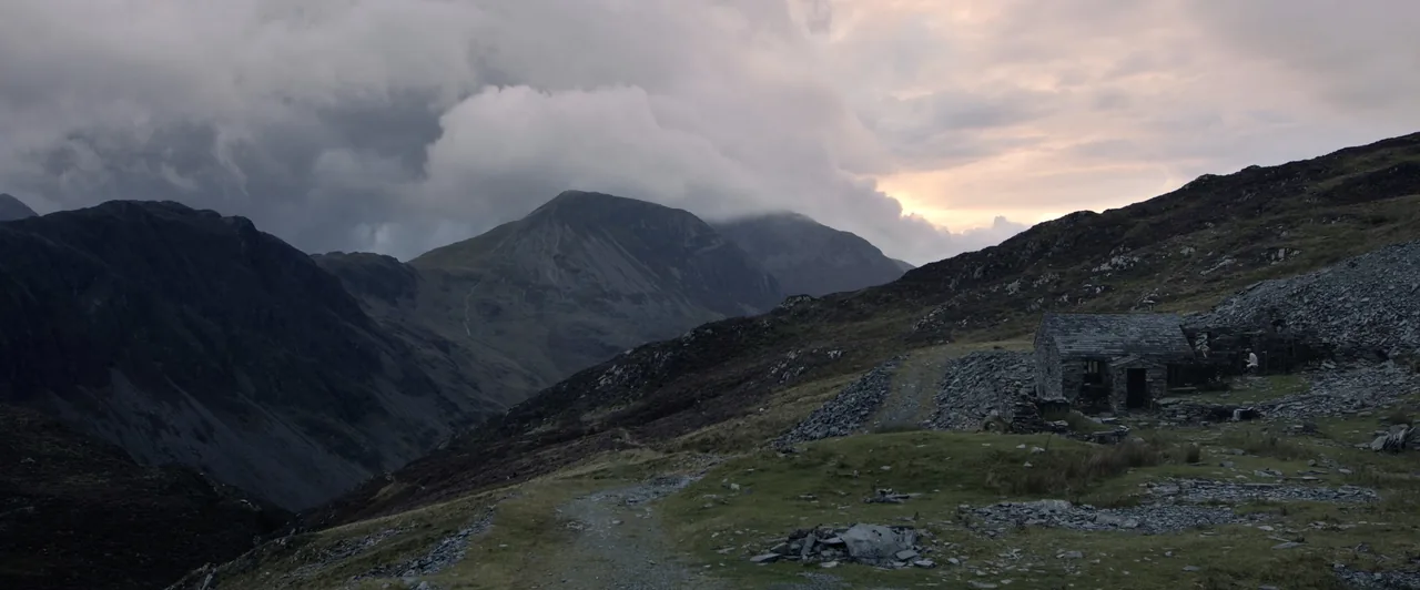 Graded still from Upper Air: the bothy on a stony fellside at dusk, cloud pouring over the high fells beyond.