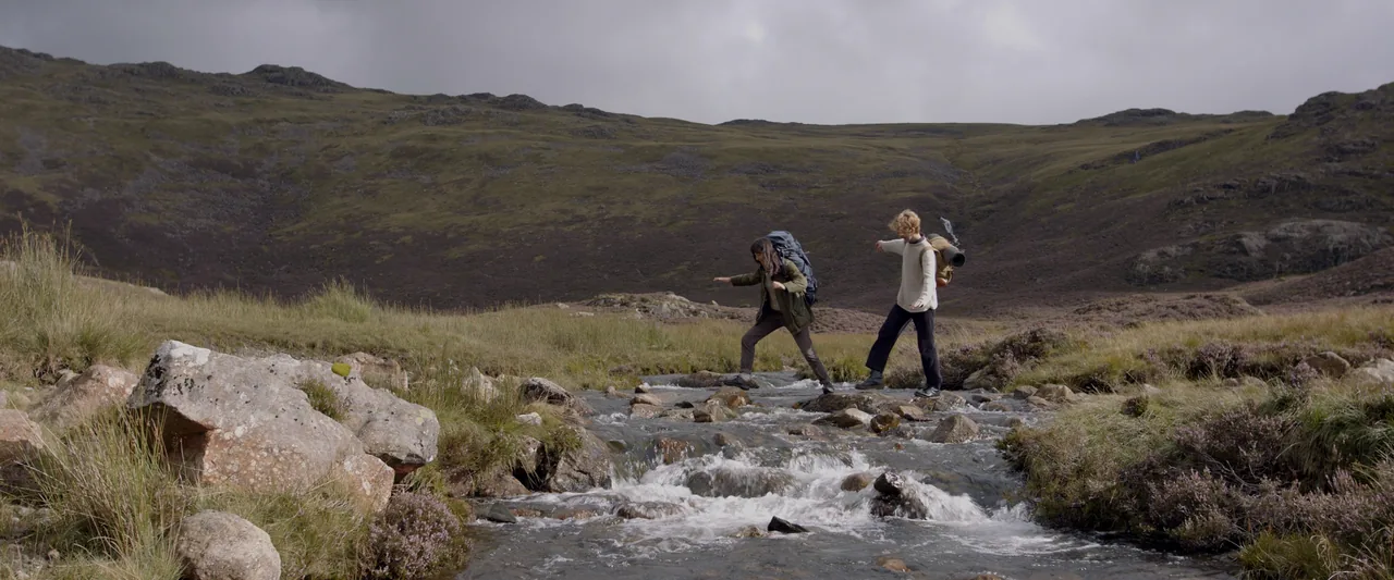 Sam and Ethan cross a rocky beck on open fellside, carrying full packs.